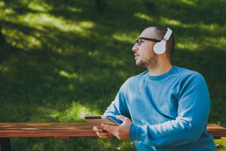 Young happy man, businessman or student in casual blue shirt glasses sitting at table with headphones, tablet pc in city park, listen music, rest outdoors on green nature. Lifestyle leisure conceptの写真素材