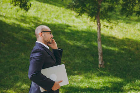 Successful smart businessman in white shirt, classic suit, glasses. Man stand with laptop pc computer, talk on mobile phone in city park outdoors on nature background. Side view, business conceptの写真素材