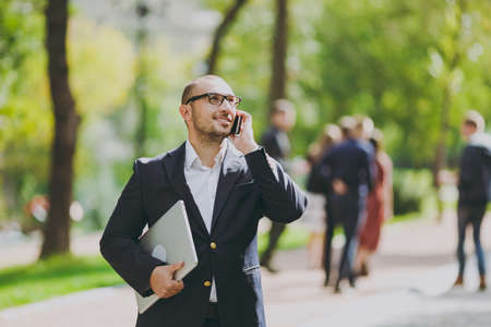 Successful smart businessman in white shirt, classic suit, glasses. Man stand with laptop pc computer, talk on mobile phone in city park outdoors on nature background. Mobile Office, business conceptの写真素材