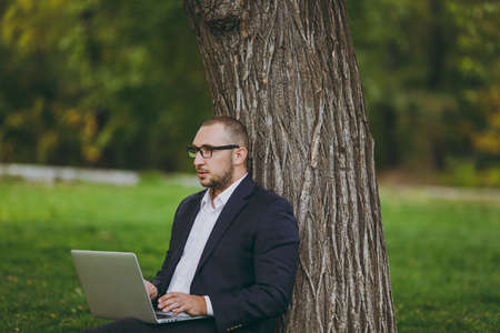 Young successful businessman in white shirt, classic suit, glasses. Man sit on grass ground, work on laptop pc computer in city park on green lawn outdoors on nature. Mobile Office, business conceptの写真素材