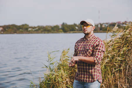 Young unshaven man in checkered shirt, cap and sunglasses looks afar and pulls out of small box...