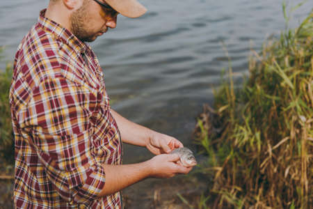 Young unshaven man in checkered shirt, cap and sunglasses caught a fish and holds it in arms on the shore of lake on background of water, shrubs and reeds. Lifestyle, recreation, leisure concept.の写真素材