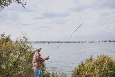 Side view Young unshaven man with a fishing rod in checkered shirt, cap and sunglasses casts a fishing pole on a lake from the shore near shrubs and reeds. Lifestyle, recreation, leisure concept.の写真素材