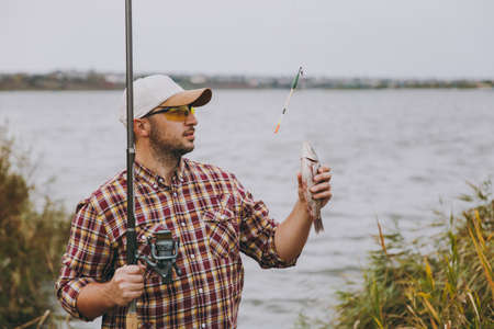 Young unshaven man in checkered shirt, cap and sunglasses pulled out a fishing pole and holds caught fish on shore of lake near shrubs and reeds. Lifestyle, recreation, leisure concept.の写真素材