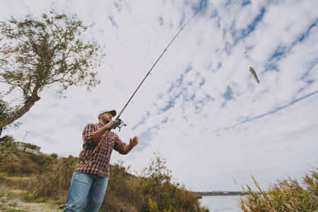 Bottom view Young unshaven man in checkered shirt, cap and sunglasses pulls out a fishing pole with caught fish on a lake from the shore near shrubs and reeds. Lifestyle, recreation, leisure concept.の写真素材