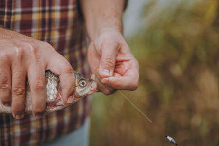 Close up Man in checkered shirt removes caught fish from a hook on a fishing rod on a blurred pastel brown background. Lifestyle, recreation, fisherman leisure concept. Copy space for advertisementの写真素材