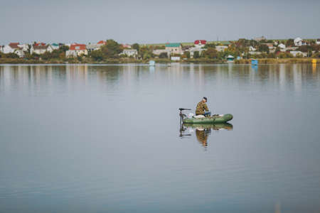 Beautiful landscape fisherman sits in camouflage jacket on rubber boat in middle of lake on houses background on sunny day. Lifestyle, recreation, man leisure concept. Copy space for advertisementの写真素材
