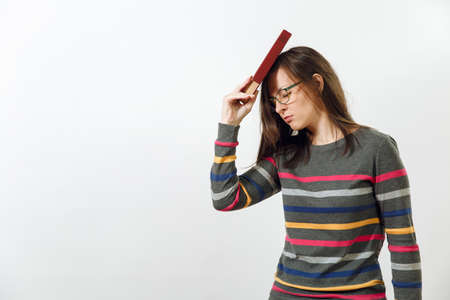 A beautiful European young brown-haired woman in glasses for sight dressed in casual dark grey longsleeve with colorful stripes, standing with book on a white background. Reading and studying conceptの写真素材