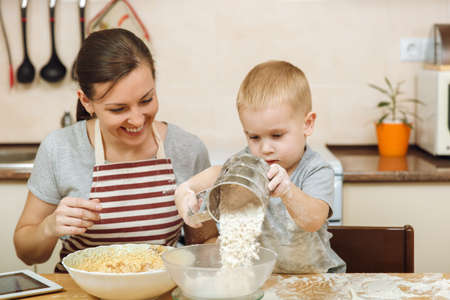 Little kid boy helps mother to cook Christmas ginger biscuit in light kitchen with tablet on the table. Happy family mom 30-35 years and child 2-3 in weekend morning at home. Relationship concept.の写真素材