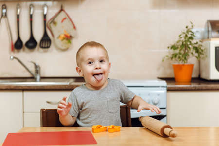 The little kid boy helps mother to cook Christmas ginger biscuit in light kitchen. Happy child in gray T-shirt 2-3 years at the table stuck out his tongue with a rolling pin and dough molds at home.の写真素材