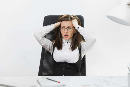 The beautiful tired perplexed and stress brown-hair business woman in suit and glasses sitting at the desk, working at contemporary computer with documents in light office on white backgroundの写真素材