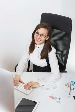 Pretty smiling brown-hair business woman in suit and glasses sitting at the desk, working at...