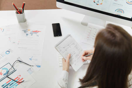 Business woman in suit sitting at the desk, writing with pencil information to notebook, working at...