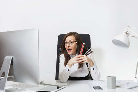 Pretty smiling brown-hair business woman in suit sitting at the desk with pencils, working at computer with modern monitor with documents in light office, looking at the camera on white backgroundの写真素材