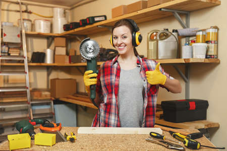 Young woman in plaid shirt gray T-shirt noise insulated headphones yellow gloves showing thumb up,...