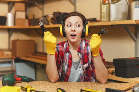 Beautiful crazy fun caucasian young woman in plaid shirt, noise insulated headphones working in carpentry workshop at table place, with two wires from electrical appliances like power drill and sawの写真素材