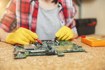 Multimeter for electrician. Close up of woman in yellow gloves, glasses digital electronic engineer repairing, soldering computer PC motherboard in workshop at wooden table with different toolsの写真素材