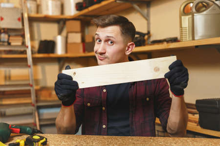 Handsome smiling caucasian young man in plaid shirt, black T-shirt, gloves working in carpentry workshop at wooden table place with piece of wood, different tools. Copy space for advertisementの写真素材