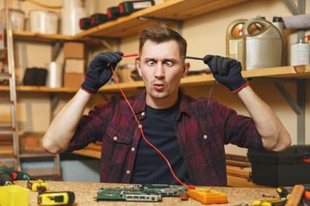 Multimeter for electrician. Crazy aggressive batty mad fun young man in plaid shirt, digital electronic engineer repairing, soldering computer PC motherboard in workshop at wooden table with toolsの写真素材