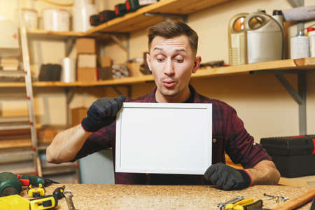 Handsome caucasian young man in plaid shirt, gloves working in carpentry workshop at wooden table place with blank frame, different tools. With empty place for text. Copy space for advertisementの写真素材