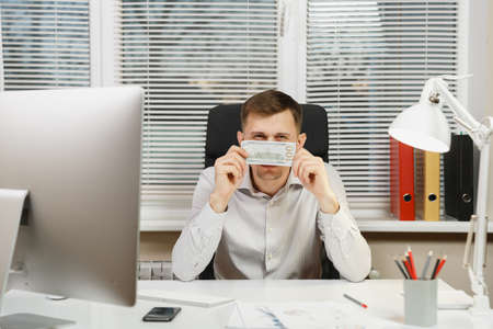 Handsome serious business man in shirt sitting at the desk hiding behind cash money, working at computer with modern monitor, documents in light office on window background. Manager or workerの写真素材