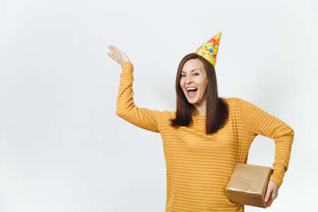 Fun young happy woman in yellow clothes, birthday party hat holding golden gift box with present, celebrating holiday, pointing hand away on white background isolated, copy space for advertisementの写真素材