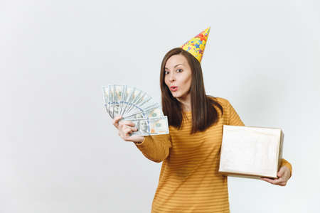 Caucasian fun young happy woman in yellow clothes, birthday party hat holding golden gift box with present and wad of cash money, celebrating holiday on white background isolated for advertisementの写真素材