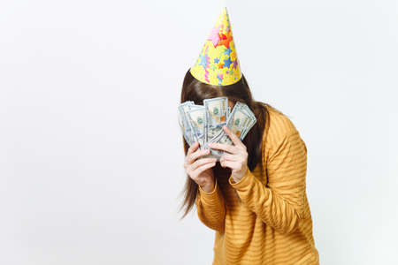 Pretty caucasian lucky young happy woman in yellow clothes, birthday party hat hiding behind wad of cash money, celebrating holiday on white background isolated for advertisement. Winner with dollarsの写真素材