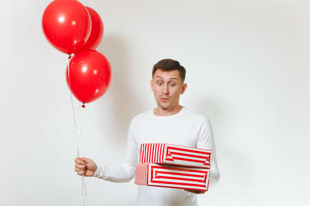 Handsome fun caucasian young happy man in longsleeve with red balloons, striped gift boxes with present, celebrating birthday, on white background isolated for advertisement. Holiday, party conceptの写真素材