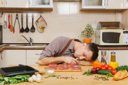 Caucasian young man in apron sleeping at table with vegetables, after cooking at home preparing meat stake from pork, beef or lamb, in light kitchen with wooden surface, full of fancy kitchenwareの写真素材