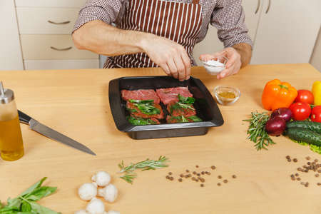 Close up young man in apron sitting at table with vegetables, cooking at home preparing meat stake from pork, beef or lamb, salt meat herbs in black baking tray, in light kitchen with wooden surfaceの写真素材