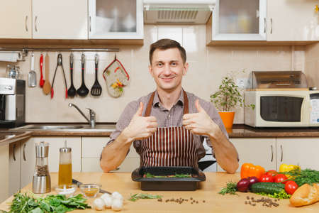 Young man in apron sitting at table with vegetables, showing thumbs up, cooking at home preparing meat stake from pork, beef or lamb, in light kitchen with wooden surface, full of fancy kitchenwareの写真素材