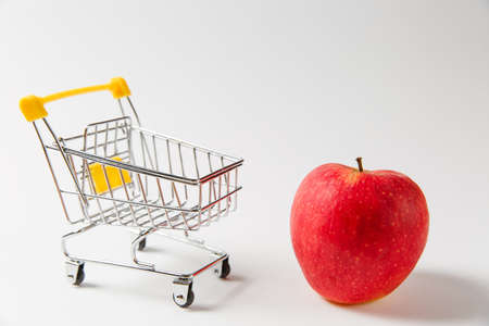 Close up of supermarket grocery push cart for shopping with yellow plastic elements on handle near bright red apple isolated on white background. Concept of shopping. Copy space for advertisement.の写真素材