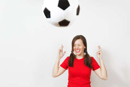 European young woman two fun pony tails, football fan or player in red uniform keep fingers crossed, flying soccer ball isolated on white background. Sport play football lifestyle concept. Make wishの写真素材