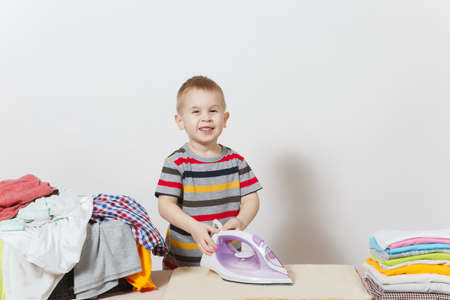 Happy little boy ironing family clothing on ironing board with iron. Kid helping with housework isolated on white background. Encouraging Autonomy in children concept. Copy space for advertisementの写真素材