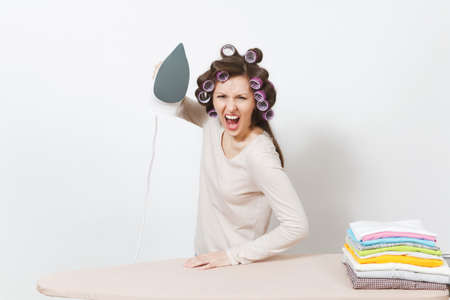 Angry screaming housewife in light clothes, curlers on hair ironing family clothing on ironing board with iron. Woman isolated on white background. Housekeeping concept. Copy space for advertisementの写真素材