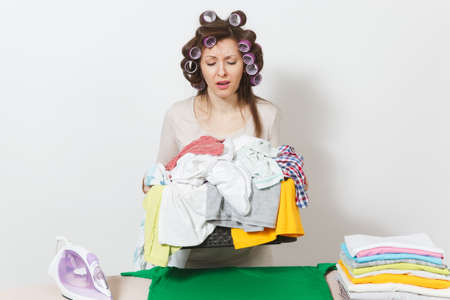 Distressed sad housewife with curlers on hair in light clothes holding basket of unshielded family clothing on ironing board with iron. Woman isolated on white background. Copy space advertisementの写真素材