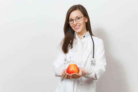 Smiling confident beautiful young doctor woman with stethoscope, glasses isolated on white background. Female doctor in medical gown holding red apple. Healthcare personnel, health, medicine conceptの写真素材