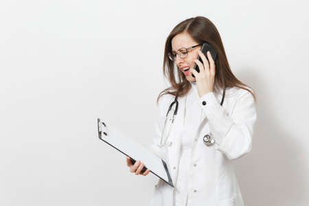 Concerned shocked doctor woman with stethoscope, isolated on white background. Female doctor in medical gown talk on mobile phone, hold health card on notepad folder. Healthcare personnel conceptの写真素材