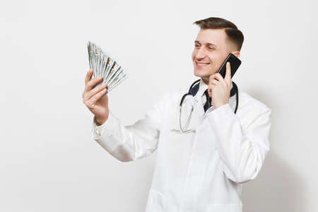 Smiling young doctor man isolated on white background. Male doctor in medical uniform talking on mobile phone, holding bundle of dollars, banknotes, cash money.の写真素材