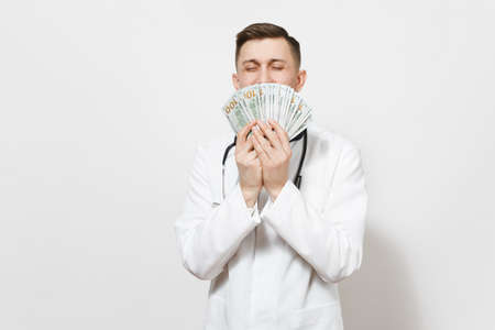 Smiling young doctor man isolated on white background. Male doctor in medical uniform, stethoscope holding bundle of dollars, banknotes, cash money. Healthcare personnel health, medicine conceptの写真素材