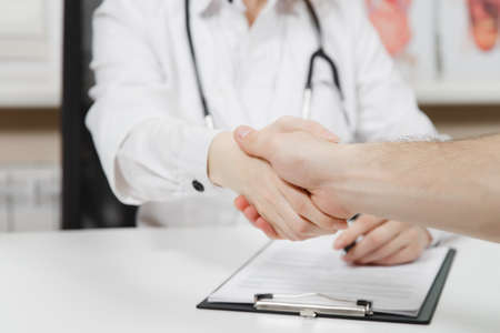 Woman sitting at desk, working with medical documents in light office in hospital. Female doctor in medical gown, stethoscope in consulting room shakes hands with patient.の写真素材