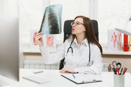 Female doctor sitting at desk, working on computer, holding X-ray of lungs, fluorography, roentgen...
