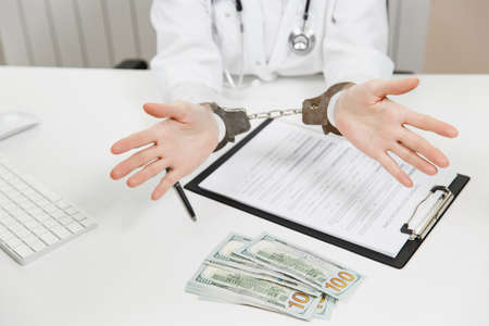 Close up arrested female doctor with medical documents at desk in hospital office. Woman in medical gown, hands with handcuffs, bundle of dollars, cash money in consulting room.の写真素材