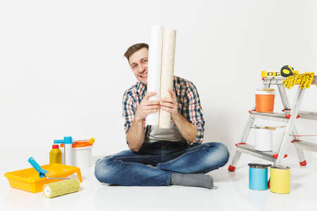 Smiling man in casual clothes sitting on floor with rolls of wallpaper, instruments for renovation apartment room isolated on white background. Gluing accessories, painting tools. Repair home conceptの写真素材