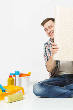 Handsome man sitting on floor with unfolded roll of wallpaper instruments for renovation apartment isolated on white background. Gluing accessories painting tools. Repair home concept. Copy spaceの写真素材