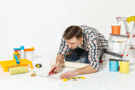 Man sitting on floor with wallpaper roll, stationery knife, instruments for renovation apartment room isolated on white background. Wallpaper, gluing accessories, painting tools. Repair home conceptの写真素材