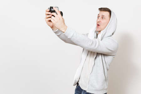 Young handsome excited student in t-shirt and light sweatshirt with hood with headphones photographs himself on retro camera in studio on white background. Concept of photography, hobby.の写真素材