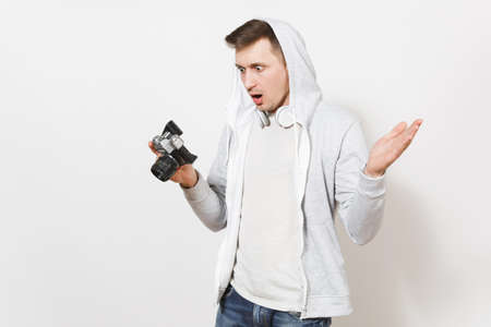 Young handsome surprised student in t-shirt and light sweatshirt with hood with headphones looks at retro camera and spreads his hands in studio on white background. Concept of photography, hobby.の写真素材