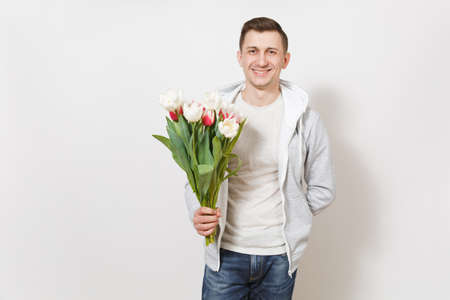 Young handsome smiling student in t-shirt and light sweatshirt holds bright bouquet of white and pink tulips in his hands in the studio on white background. Concept of celebration, good mood.の写真素材